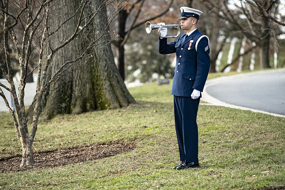 Musicien en uniforme bleu jouant du clairon, debout sur une pelouse de cimetière lors d'une cérémonie commémorative